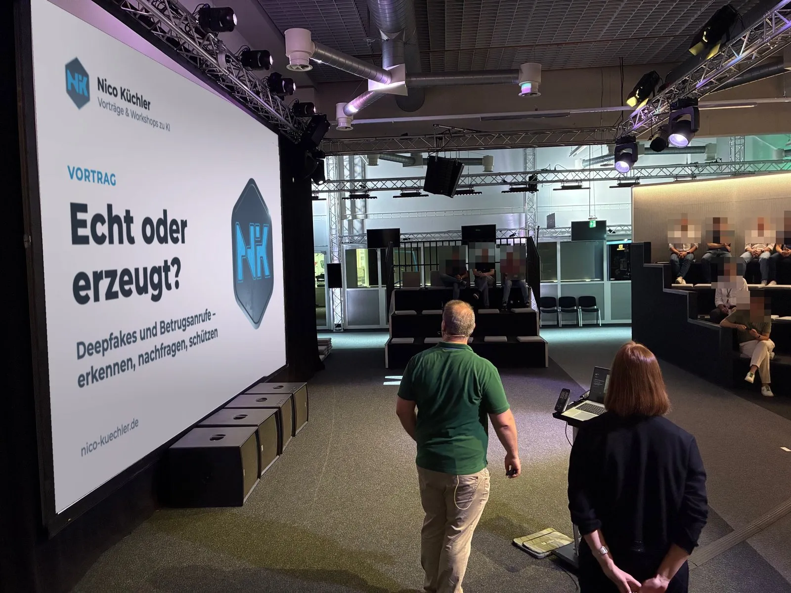 Nico Küchler auf der Bühne geht zur Leinwand mit der Folie „Echt oder erzeugt? Deepfakes und Betrugsanrufe“, das Publikum ist verpixelt